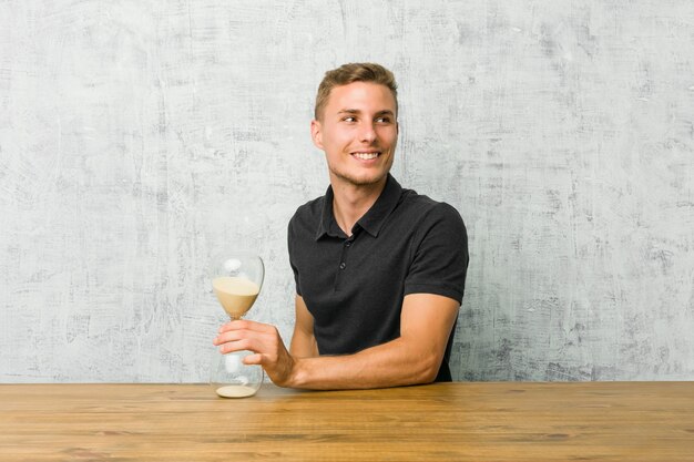 Young man holding a sand timer on a table looks aside smiling, cheerful and pleasant.