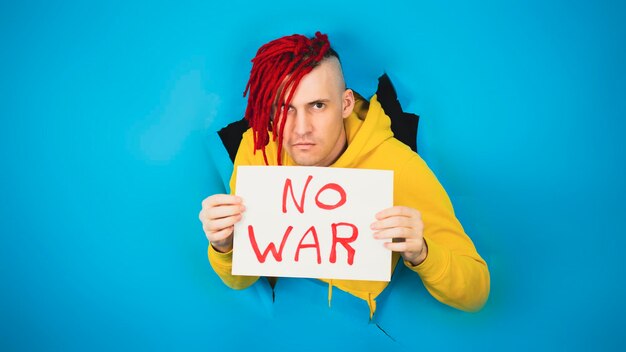 Young man holding sheet of paper No War Serious male in yellow sweatshirt sticking out of hole of blue background meaning Ukrainian flag Concept of peaceful protest