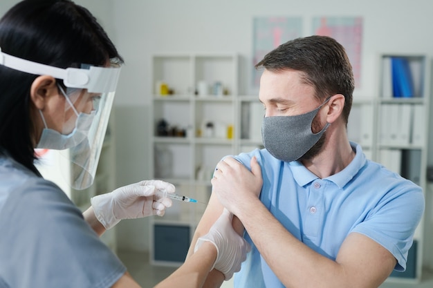 Young man holding sleeve of his shirt while female clinician in protective workwear making him injection during vaccination against covid