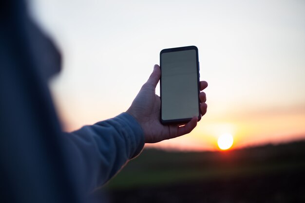 Young man holding smartphone with empty white mockup on background of sunset