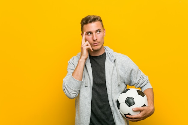 Young man holding a soccer ball pointing his temple with finger, thinking, focused on a task