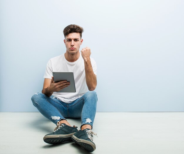 Young man holding a tablet sitting indoor showing fist, aggressive facial expression.