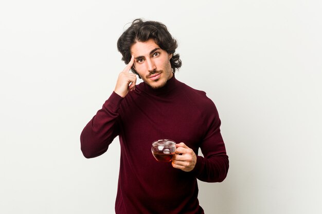 Young man holding a tea cup pointing temple with finger, thinking, focused on a task.