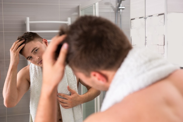 Young man holding towel on his shoulders after washing procedures while correcting hair standing in the modern tiled bathroom