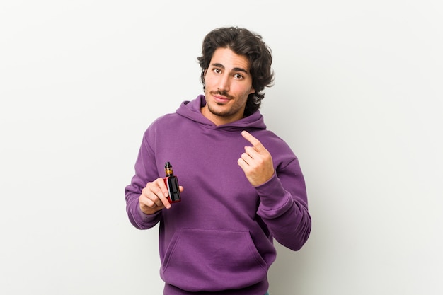 Young man holding a vaporizer pointing with finger at you as if inviting come closer.