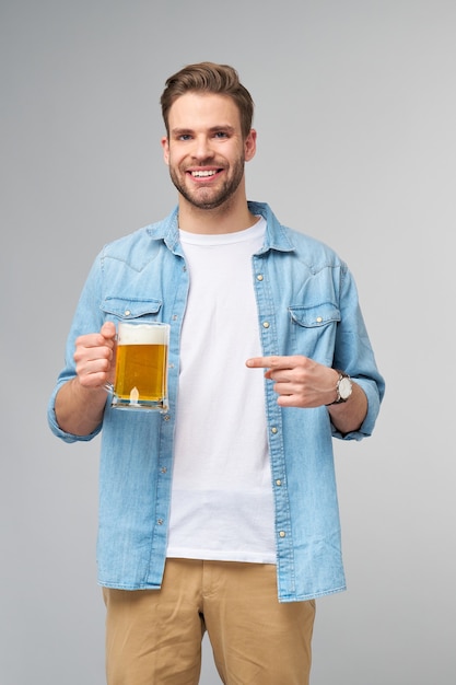 Giovane uomo azienda indossa jeans shirt tenendo un bicchiere di birra in piedi sopra il muro grigio.