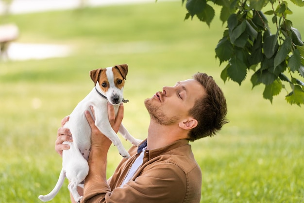 Young man holding white jack russell terrier dog with brown spots on head near tree branch