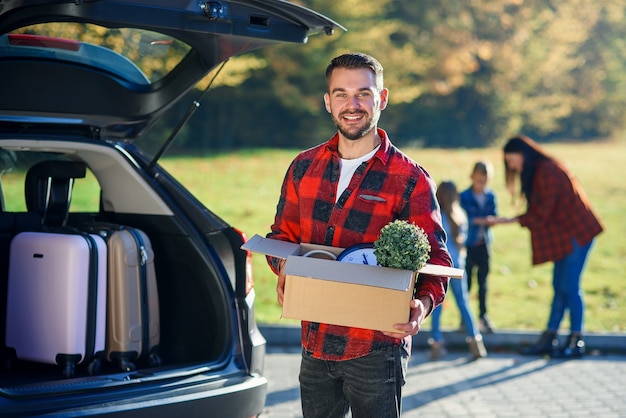 A young man holds cardboard box unloading luggage from a family\
car after moving to a new home.