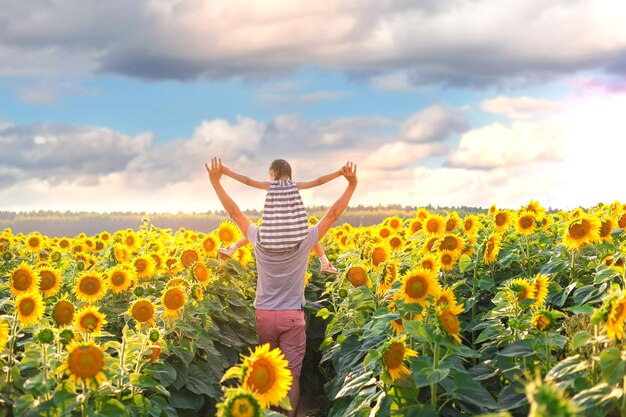 Young man holds a child on his shoulders and points with his hand to something on a sunflower field