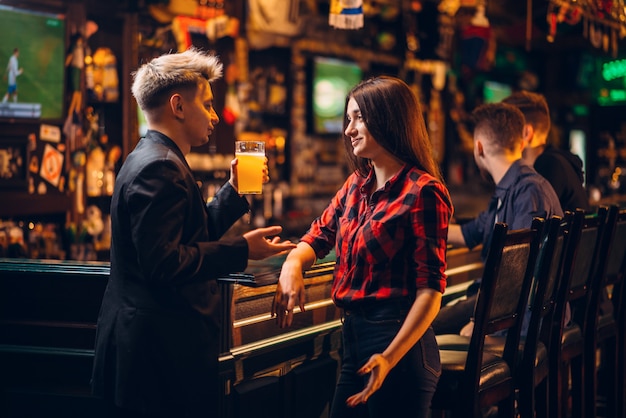 Young man holds glass of beer in hand and talks with woman at the bar counter in a sport pub, happy leisure of football fans