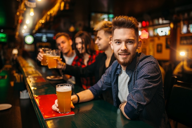 Il giovane tiene il vetro con la birra al bancone del bar in un pub sportivo, tifosi di calcio felici sullo sfondo