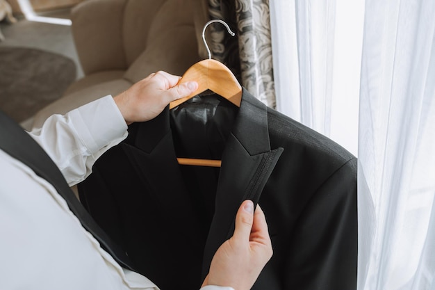 Photo a young man holds a jacket in his hands in his room near the window in a hotel room closeup photo the groom is preparing for the wedding ceremony