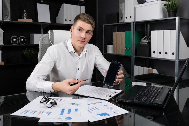 Young man holds pen in his hand and points her to phone screen sitting at table in bright office