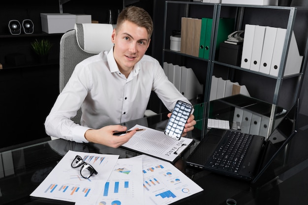 Young man holds pen in his hand and points her to phone screen sitting at table in bright office