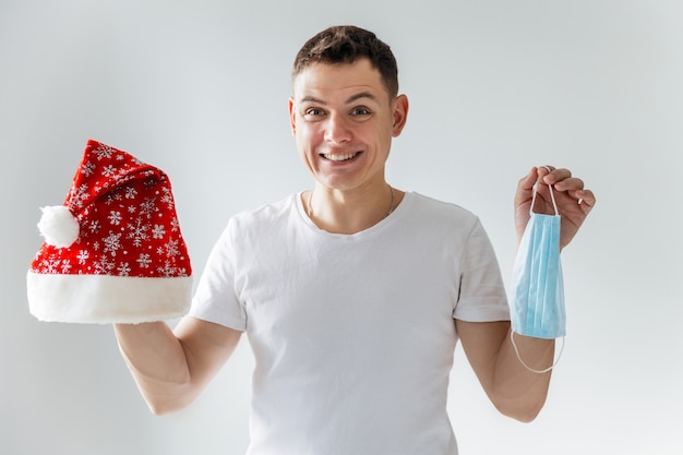 Young man holds a Santa hat and a protective medical mask in her hands