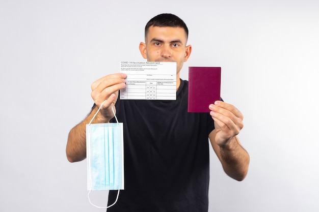 Young man holds a vaccination card passport and a medical mask on a white isolated background