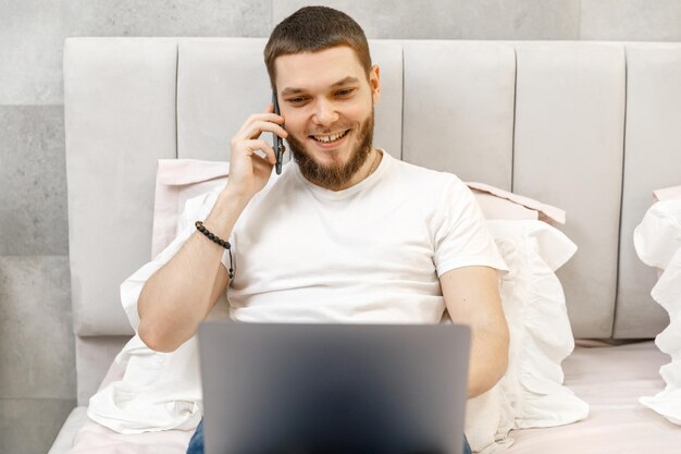 Молодой человек дома на диване разговаривает по телефону и смотрит на ноутбук