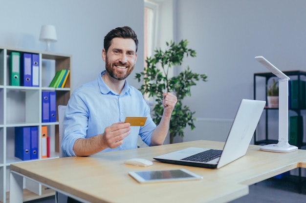 A young man at home looks at the camera and smiles holding a credit bank card for online shopping in the store rejoices a successful buyer uses a laptop