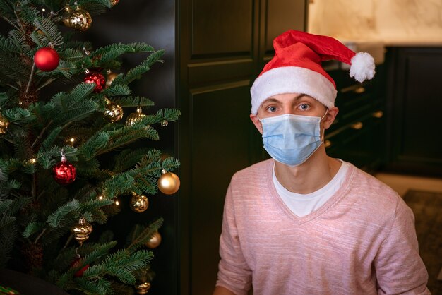 Young man at home near a christmas tree wearing a mask and a santa claus hat looks at the camera