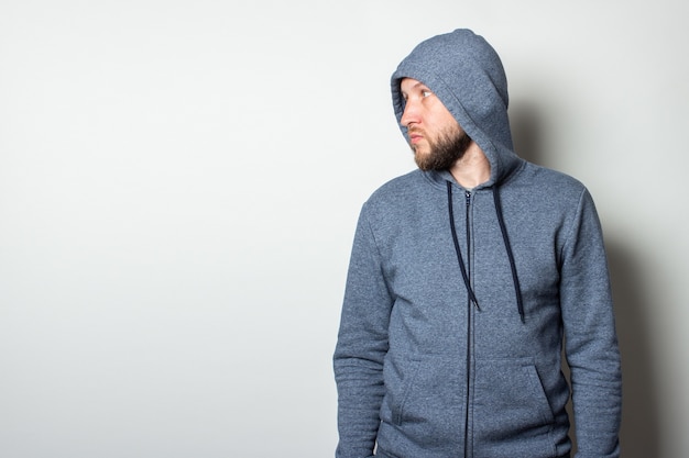 Young man in a hood with a serious, aggressive face looks to the side against a light wall