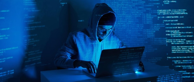 暗い背景にコンピューターを使用するフードシャツを着た若い男性