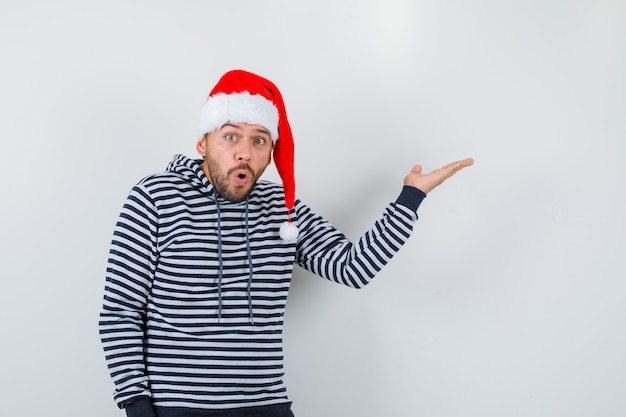 Young man in hoodie, Santa Claus hat pretending to show something and looking wondered , front view.