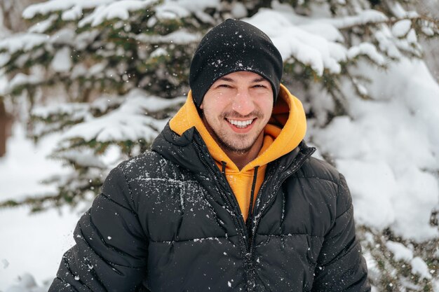 Young man in hoodie and warm jacket standing in a snowy park and smiling