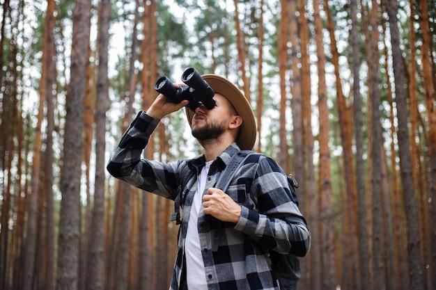 Фото Молодой человек в шляпе с рюкзаком и биноклем в сосновом лесу поход в горы или лес