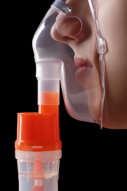 Young man inhaling through respiratory inhaler mask on black background. Use nebulizer for inhalation therapy for asthma and respiratory diseases. Side view of nose and lips in mask.