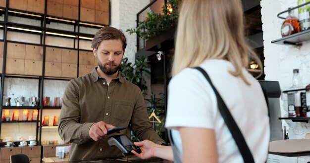 若い男が喫茶店で持ち帰り用のコーヒーを購入し、スマートフォンで非接触型決済を行っています。現代のテクノロジーと銀行のコンセプト。