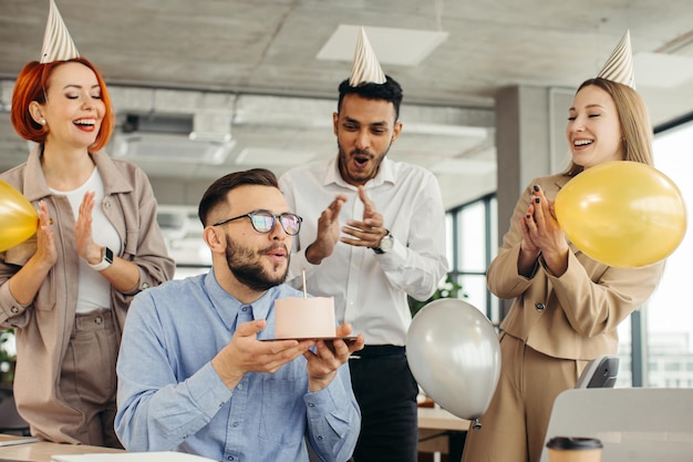 若い男はケーキにろうそくを吹き、同僚と誕生日を祝いながら願い事をしようとしています オフィスで誕生日を祝う同僚