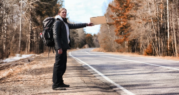 Photo a young man is hitchhiking around the country. the man is trying to catch a passing car for traveling. the man with the backpack went hitchhiking to south.