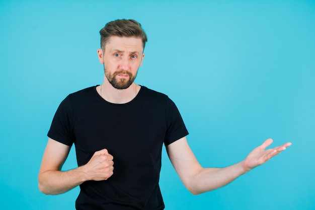 Young man is looking at camera by pointing rigth with hand on blue background