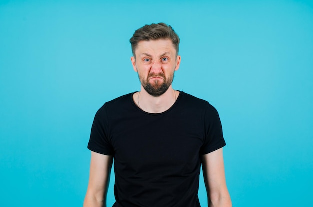 Young man is looking at camera by showing angry mimicry on blue background