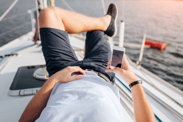 Young man is lying on yacht board and chilling. he holds phone\
in lefy hand. right one is on stomach. he keeps right leg on left.\
man has vacation. he wears white shirt and black shorts with\
shoes