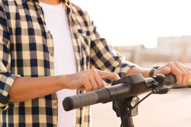 Young man is pushing start button of modern gadget and driving along street of city. Closeup male hands on wheel of electric scooter. Ecological alternative transport concept