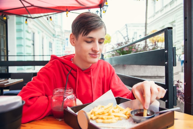 Молодой человек сидит в уличном кафе и ест картофель-фри.
