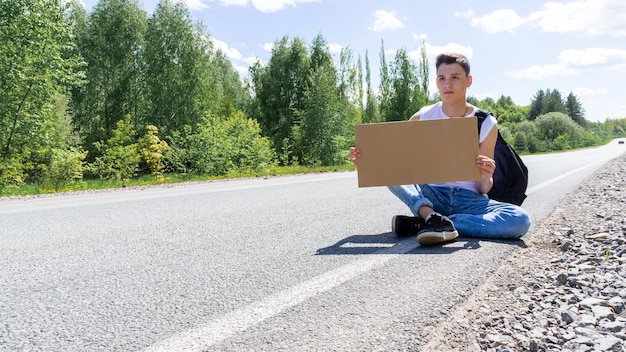 A young man is sitting on the side of the road with a cardboard sign in his hands Space for text