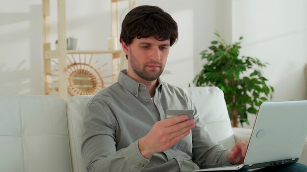 Young man is sitting on the sofa in the living room, making online purchases through a laptop, holding a credit card, paying for purchases in an online store resting at home