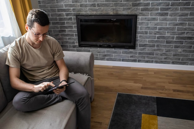 A young man is sitting on a sofa and playing on a tablet A guy is typing something Online job on a tablet device