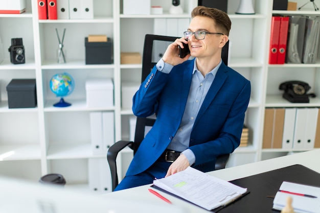 A young man is sitting at a table in the office and talking on the phone.