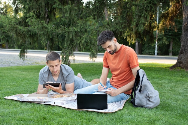 Young man is sitting with a laptop and tablet on grass on a college campus and taking notes in notebook.