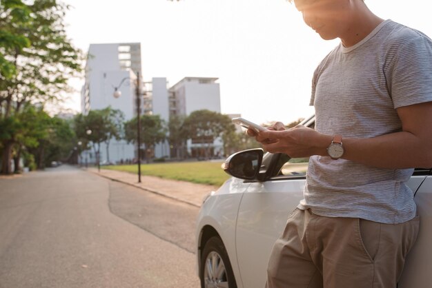 若い男が電気自動車の近くに立って、スマートフォンを見ています。レンタカーは電気自動車の充電ステーションで充電しています。カーシェアリング。