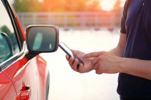 Молодой человек стоит возле своей машины с помощью смартфона на закате