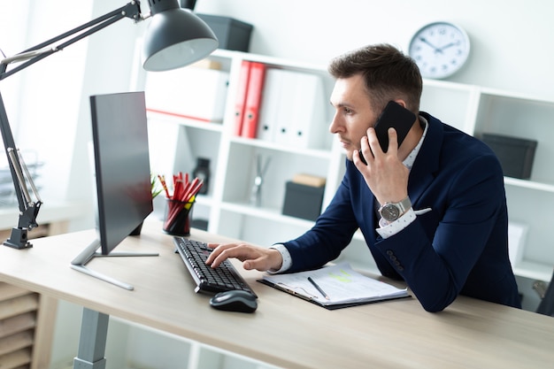 A young man is standing near a table in the office, talking on the phone and typing text on the keyboard.