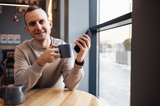 若い男性がバーで携帯電話からテキストメッセージを送信し、カプチーノを飲んでいます。昼食と仕事中にシティカフェでエスプレッソコーヒーを飲む若いファッショナブルな男