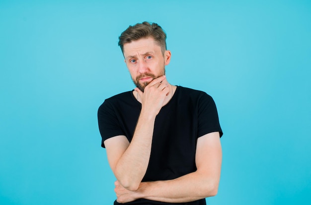 Young man is thinking by holding hand on chin on blue background