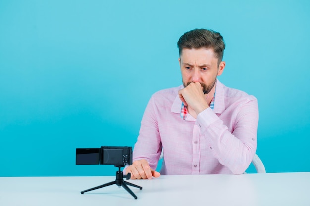 Young man is thinking by holding hand on chin and sitting in front of his mini camera on blue background