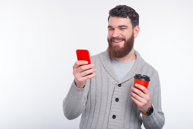 Young man is tying a message on his phone and holding a red cup to go on white background.