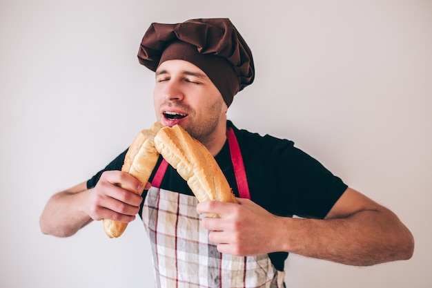 Giovane uomo isolato su sfondo. ragazzo in divisa da cuoco che morde due pezzi di baguette francese e lo assapora. mangiando pane francese delizioso saporito fresco.